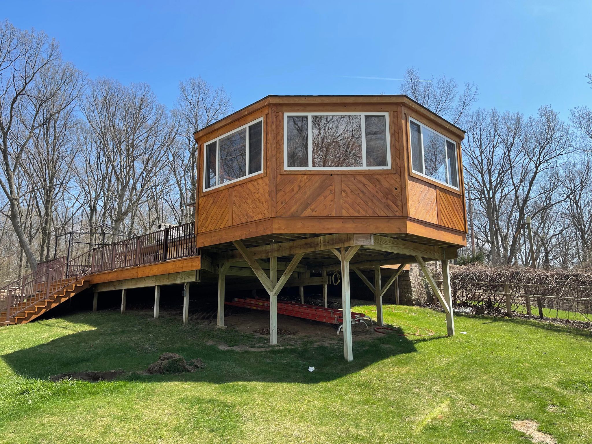 Elevated wooden deck and sunroom on stilts with fresh wood staining, surrounded by green lawn and leafless trees, showcasing professional exterior deck staining and pressure washing services in Flint, MI